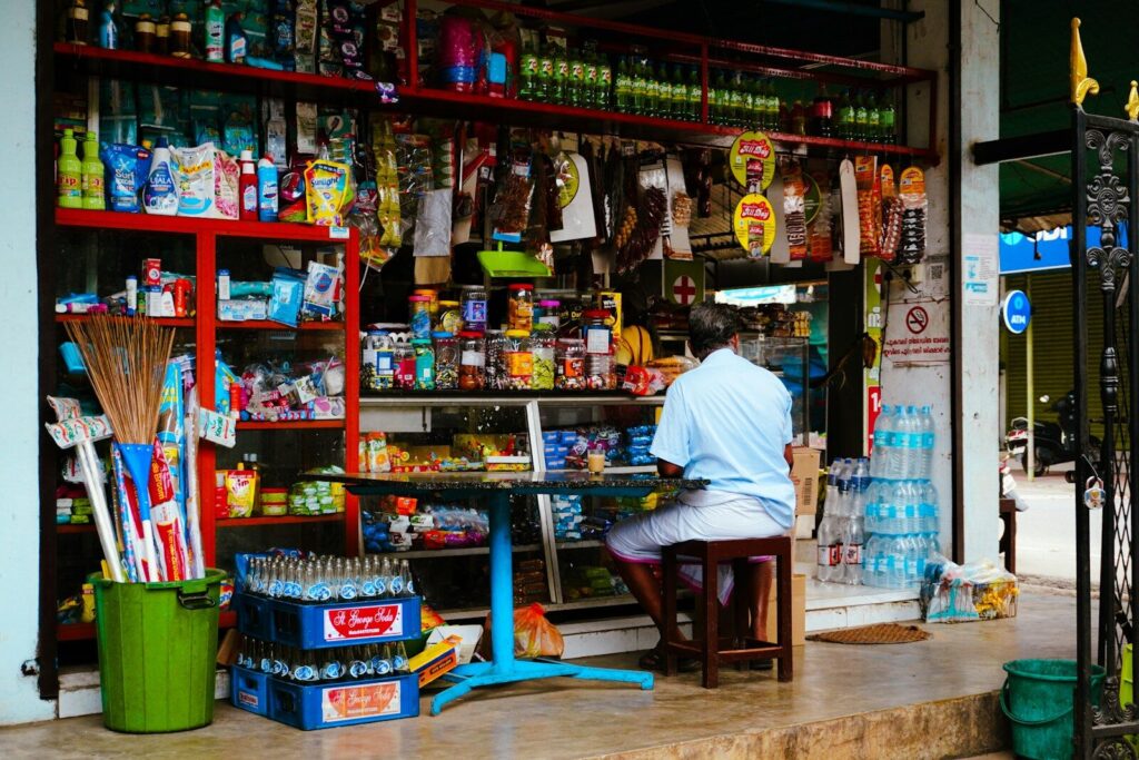 Man sits at a small table outside a well-stocked store.
